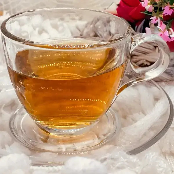 A clear glass cup filled with a warm, golden brew of Jaroma's Happy Morning Earl Grey tea, resting on a saucer. The tea has a light amber hue, with delicate reflections on the surface. The background features soft, neutral tones, creating a calming and inviting atmosphere for a morning tea experience.