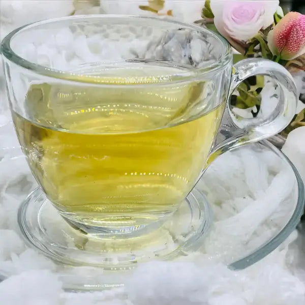 A clear glass teacup filled with a golden infusion of Jaroma Goddess Bliss Tea. The soothing blend, surrounded by delicate floral accents, exudes a calming ambiance. The tea's vibrant yellow color hints at the nourishing properties of organic ingredients like burdock root, peppermint, and calendula petals, ideal for wellness and relaxation.